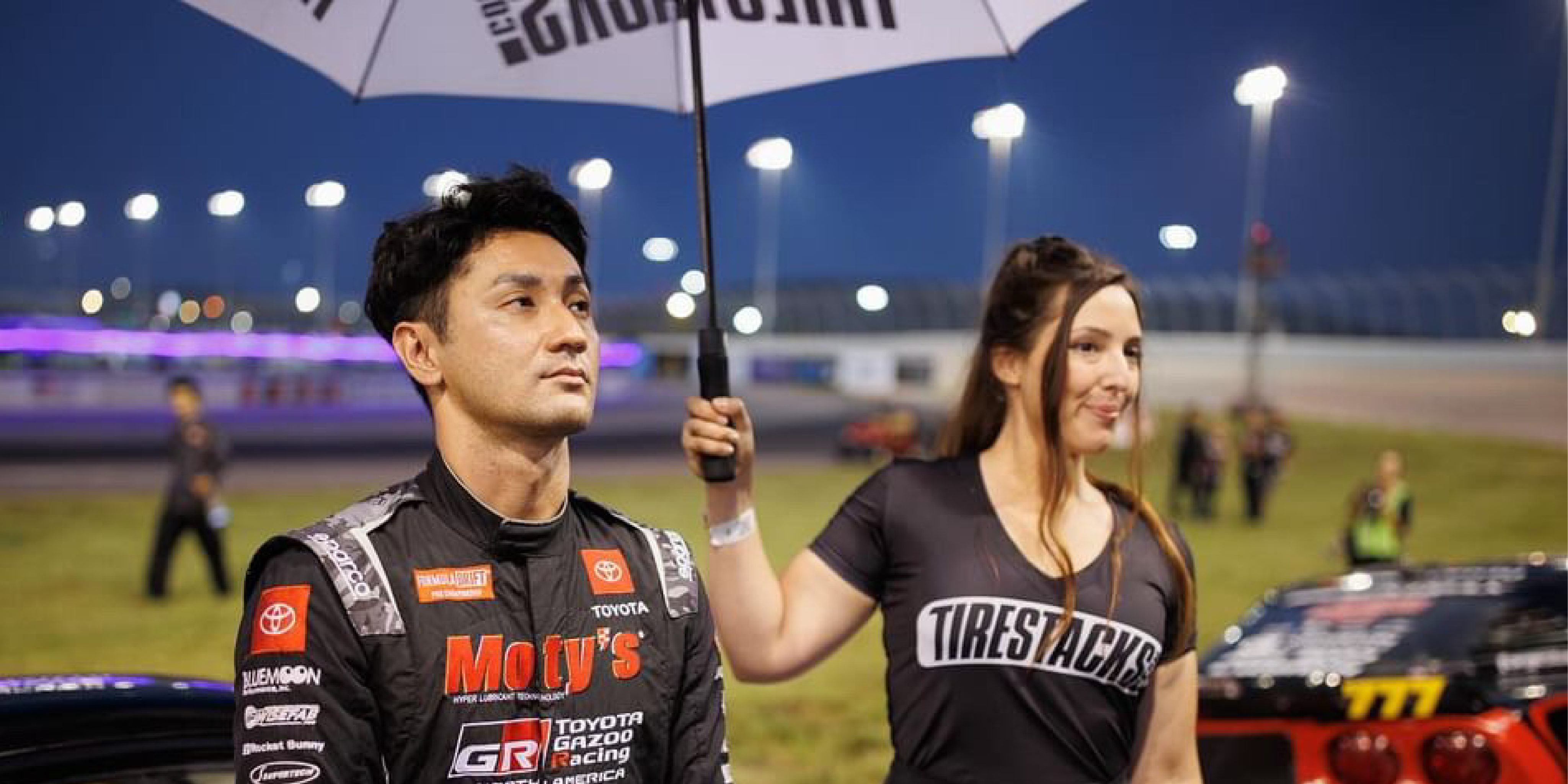 Ken Gushi, a Formula D driver, stands next to a woman holding an umbrella at a nighttime racing event with bright  lights.
